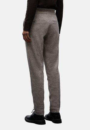 Osoba v šedých kostkovaných kalhotách slim-fit, černých šněrovacích botách a černém dlouhém rukávu, stojící na jednobarevném pozadí.