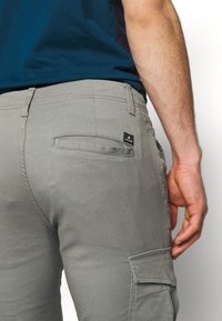 Main d'homme reposant à côté de la poche d'un pantalon cargo gris, portant une chemise bleu foncé avec une poche arrière visible et des passants de ceinture.