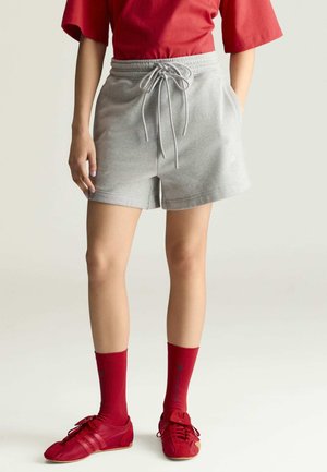 Persona con pantalones cortos grises con cordón, calcetines rojos hasta el tobillo, zapatillas deportivas rojas y camiseta roja de manga corta con las manos visibles en los bolsillos.