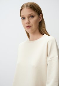 Felpa beige chiaro con collo a giro, vestibilità rilassata e texture morbida. Presenta un colletto a costine e maniche leggermente oversized.