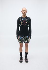 Mann står foran og har på seg svart langermet skjorte, shorts med blomstertrykk, matchende blomstervest, svarte sokker og svarte joggesko.