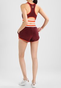 Sujetador deportivo burdeos con detalles en naranja y diseño de espalda cruzada, combinado con pantalones cortos burdeos con rayas blancas y naranjas, usados con zapatillas blancas.