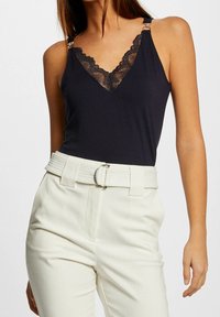 Camisole noire avec dentelle et accents en métal argenté, associée à un pantalon blanc taille haute doté d'une ceinture en tissu avec une boucle en métal.