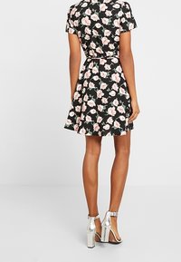 Kvinna som bär en svart blommig kortärmad klänning och silverfärgade högklackade sandaler, stående vänd bort från kameran mot en enfärgad bakgrund.