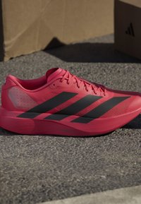 Κόκκινο παπούτσι τρεξίματος Adidas με μαύρες ρίγες τοποθετημένο σε τραχύ γκρι πλακόστρωτο κοντά σε χαρτοκιβώτια.
