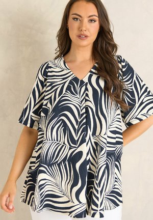 Kobieta z długimi brązowymi włosami, ubrana w luźną bluzkę z dekoltem w kształcie litery V, wzór tropikalnych liści w kolorze czarno-białym, z krótkimi rękawami, na jednolitym tle.