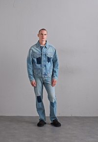 Férfi áll egy sima falnak támaszkodva, világoskék farmerdzsekit és farmert viselve, sötétkék szív- és négyzetmintás foltokkal, fekete cipővel.