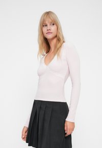 Maglietta a maniche lunghe rosa chiaro con scollo a V e bordo in pizzo, abbinata a una gonna nera plissettata. Il tessuto appare morbido e leggermente rigato.