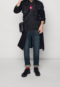 Trinchera negra sobre una sudadera con capucha negra con un logo rojo, combinada con jeans oscuros y zapatillas negras. Incluye un pequeño bolso cruzado negro.