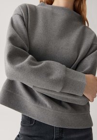 Felpa grigia in fleece con scollatura rotonda, polsini e orlo a coste. Presenta una vestibilità comoda e una texture morbida. Indossata con jeans scuri.