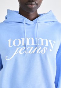 Haut à capuche bleu clair en tissu doux, arborant un logo "tommy jeans" blanc et bien visible dans une police élégante, avec des cordons de serrage ajustables.