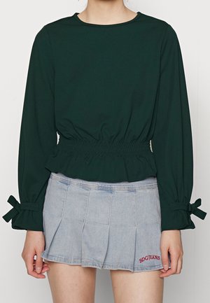 Person wears a dark green long-sleeve top with cinched waist and bow details at cuffs, paired with a light blue pleated mini skirt.