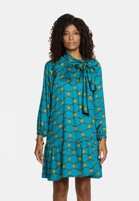 Turkusowa sukienka z długimi rękawami, zebranym dekoltem, przewiązaną kokardą i rozkloszowanym dołem. Ozdobiona żółtymi i brązowymi wzorami geometrycznymi.