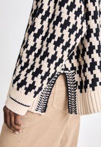 Strickpullover im beige-navy Hahnentritt-Muster, mit gerippten Bündchen und Saum, seitlichen Schlitzen und strukturiertem Stoff. Kombiniert mit beigen Hosen.