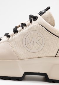 Sneaker beige con un upper in tela testurizzata, dettagli in pelle liscia, lacci neri e un prominente logo MK in rilievo sul lato.