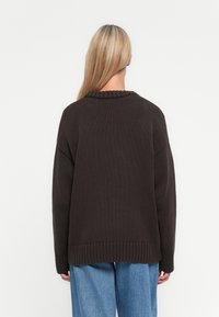 En blond person som vender bort, iført en mørk brun strikkegenser og løse blå jeans foran en enkel hvit bakgrunn.