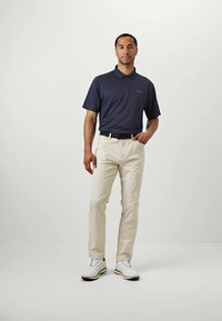 Bărbat îmbrăcat cu un tricou polo bleumarin, pantaloni bej, curea neagră și încălțăminte albă, stând pe un fundal alb simplu.