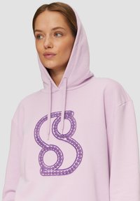 Felpa lilla con un grande logo ricamato viola, dotata di coulisse e una texture morbida. Design semplice e dal taglio rilassato.