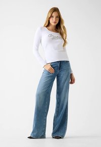 Haut à manches longues blanc avec texte gris brodé, associé à un jean large bleu clair, présentant une texture denim classique.