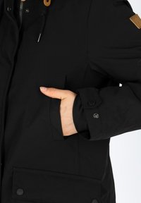 Black jacket featuring a front pocket, adjustable cuffs with snap closures, and a drawstring hood. Smooth texture and contemporary design.