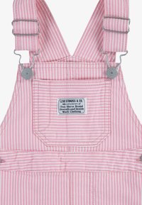 Salopette rayée rose et blanche en tissu avec des clips en métal. Comprend une poche avant avec une étiquette indiquant la marque et le fabricant.