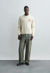 Krém színű pamut pulóver bal mellen texturált folttal, világoszöld, széles szárú farmernadrággal és fekete cipővel párosítva.