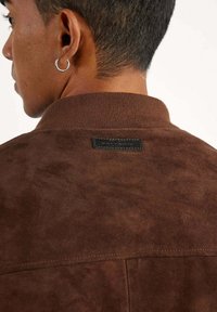 Veste en suède marron avec col côtelé, coutures visibles et une petite étiquette noire au dos. Texture lisse avec une finition mate.