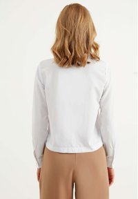 Witte lange mouwen shirt met een kraag en detail op de achterkant, gecombineerd met beige high-waisted broeken. De stof lijkt glad en lichtgewicht te zijn.