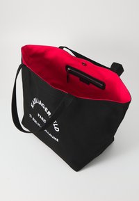 Sac fourre-tout noir en toile avec un intérieur rose vif, présentant une poche zippée et un texte imprimé blanc sur un côté. Deux anses robustes.