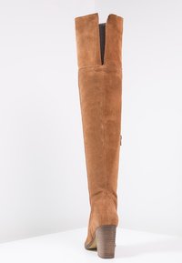 Alto stivale in suede marrone con tacco a blocco in legno sovrapposto e fessura posteriore in alto, su una superficie bianca contro uno sfondo bianco.