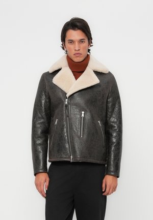 Veste en cuir marron avec un col en shearling crème, poches zippées et finition texturée. Portée sur un pull col roulé marron.