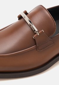 Hnědé kožené mokasíny s hladkým povrchem, vybavené stříbrnými kovovými detaily a prošitým podpatkem. Okrúhlá špička a elegantní design.