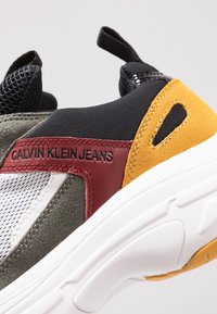 La sneaker Calvin Klein Jeans présente un mélange de suède gris, de maille noire et d'accents en cuir rouge, avec une épaisse semelle blanche et un détail jaune.