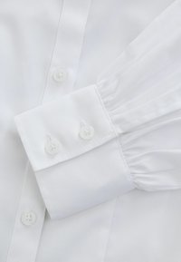 Camicia bianca con polsini button-down e dettagli plissettati. Il tessuto è liscio e presenta una leggera lucentezza, mentre i bottoni sono in plastica bianca.