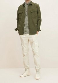 Camisa overshirt de pana verde oliva con dos bolsillos en el pecho, superpuesta a una camiseta gris claro, combinada con pantalones cargo blancos y zapatillas blancas.