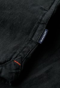 Tessuto nero con una consistenza liscia. Presenta un'etichetta blu con "Superdry" e un piccolo dettaglio di cucitura arancione sul bordo.