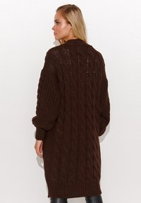 Brązowy sweter z dzianiny z wzorem w warkocze, długimi rękawami i luźnym krojem, sięgający do połowy ud z lekko oversized sylwetką.