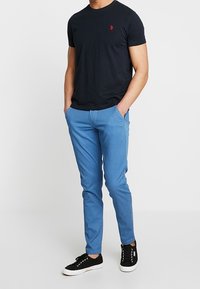 Mörkblå bomullst-shirt med en liten röd logotyp, kombinerad med ljusblå texturerade chinos och svarta sneakers med vita sulor.