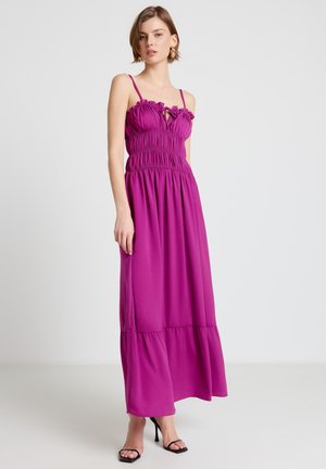 Donna che indossa un abito lungo senza maniche di un vivace viola, con corpetto arricciato e spalline sottili, abbinato a sandali neri con tacco alto.