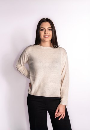 Beżowy sweter dzianinowy z okrągłym dekoltem i prążkowanymi wykończeniami u dołu i na mankietach. Model nosi czarne spodnie, tło jest jednolicie białe.