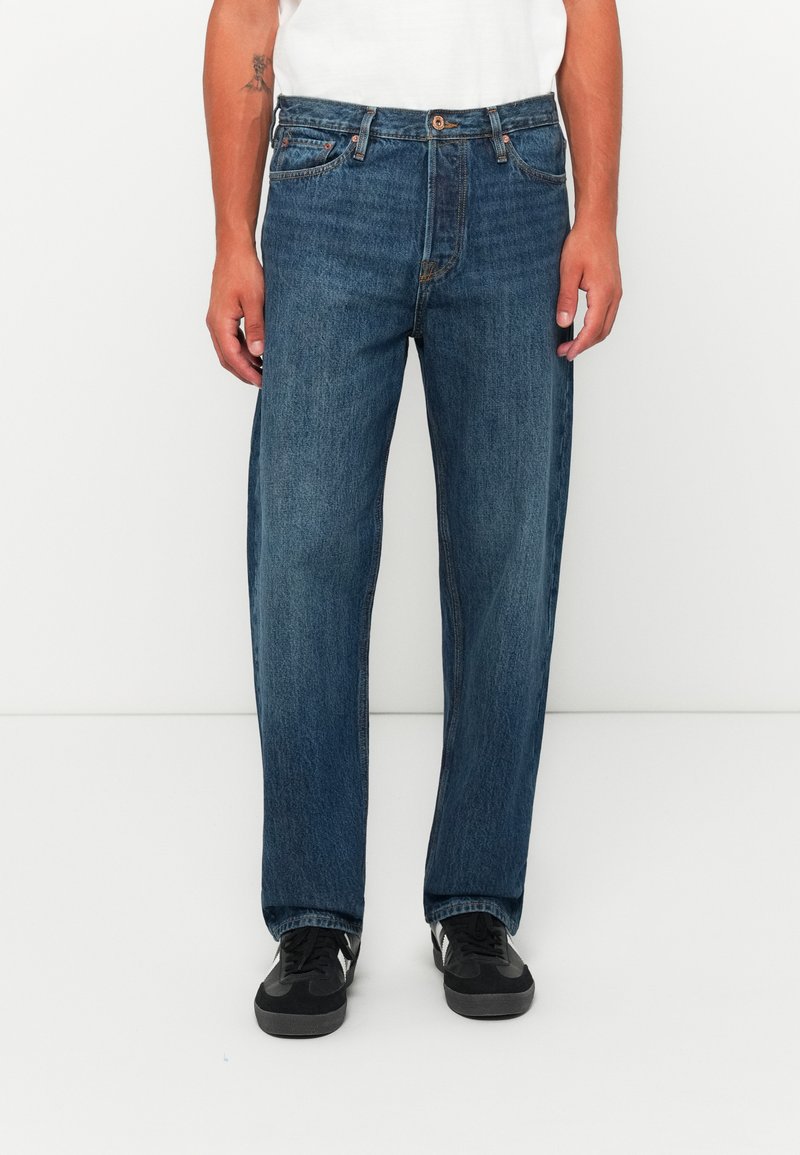 Mid-wash straight-leg jeans worn with black sneakers and a white top, hands relaxed by the sides against a plain white background.