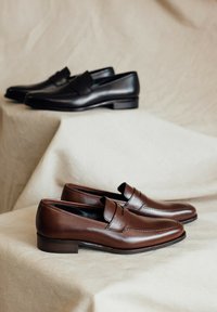 Bruine leren instappers met een gladde afwerking, gestikte accenten en een lage hak. Zwarte leren instappers met een gepolijste oppervlakte staan erachter.