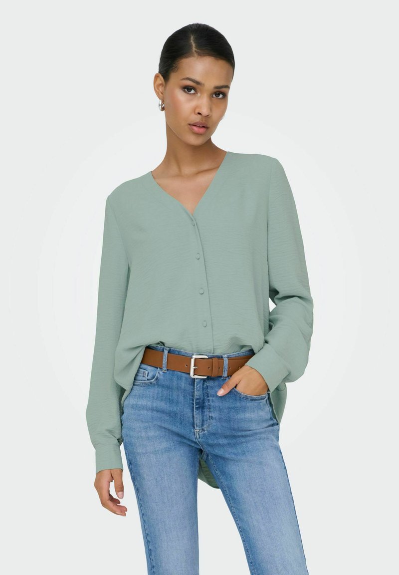 Femme portant un chemisier à manches longues vert clair boutonné, rentré dans un jean bleu avec une ceinture marron, posant avec une main dans la poche.