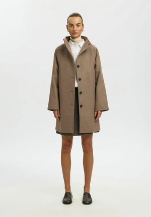 Femme debout portant un manteau marron boutonné sur une chemise blanche et une jupe foncée avec des chaussures noires sans lacets sur un fond uni.