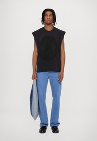 Melnas bezpiedurkņu blūzes ar teksturētu logotipa dizainu, apvienota ar gaiši ziliem džinsiem un melniem apaviem, turam pelēku zilo džinsu jaku.