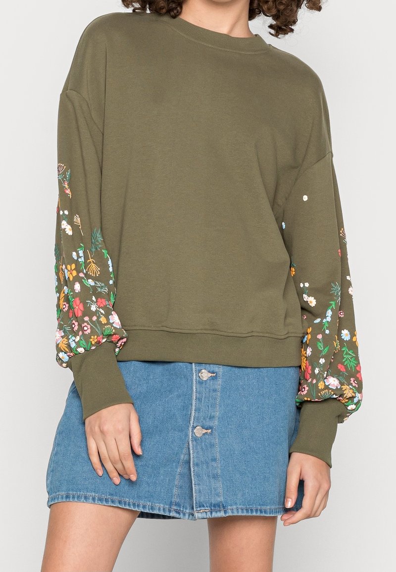 Personne portant un sweat-shirt vert olive avec des manches à imprimé floral et une jupe en jean bleue à boutons sur le devant.