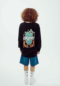 Schwarzes Langarmshirt mit einem bunten Grafikdesign, das "GRIMY" zeigt, kombiniert mit blauen Jeansshorts und weißen Socken über weißen Schuhen.