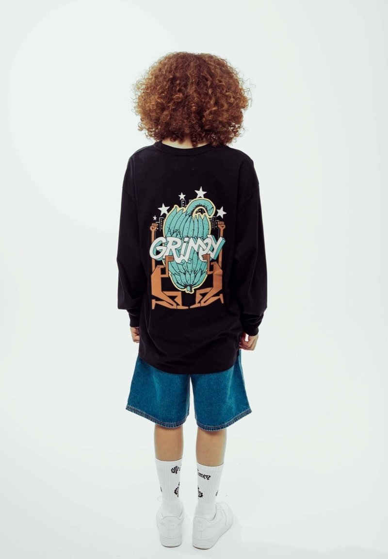 Schwarzes Langarmshirt mit einem bunten Grafikdesign, das "GRIMY" zeigt, kombiniert mit blauen Jeansshorts und weißen Socken über weißen Schuhen.