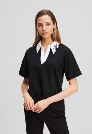 Haut noir à manches courtes avec un col blanc pointu arborant un design signature. Le matériau semble lisse et décontracté.