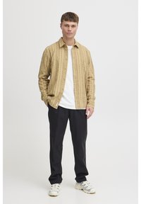 Light brown, patterned button-up shirt over a white t-shirt, paired with black pants and white sneakers. Model stands against a plain background.